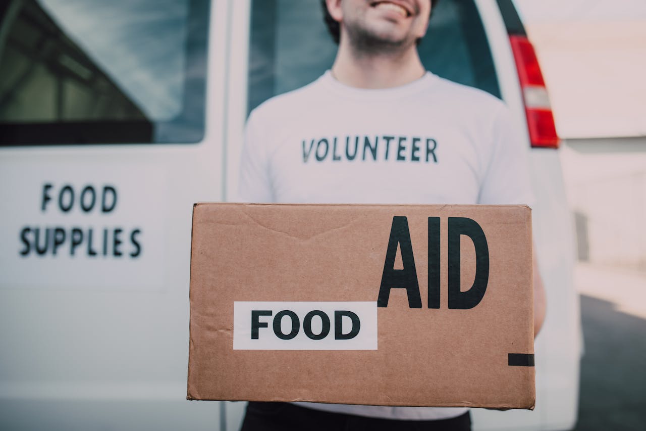 services-03 Smiling volunteer holding a box labeled 'Food Aid' outside a van with 'Food Supplies' sign.